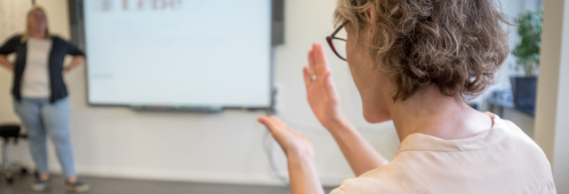 Et underviser står med hænderne i siden i baggrunden og ser på en kursist, som siger noget på tegnsprog